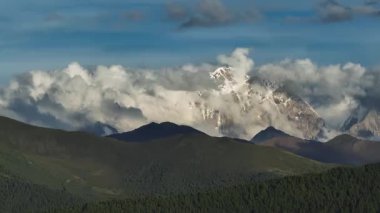 Mavi gökyüzünün altında bulutlu görkemli kar örtülü dağlar. 