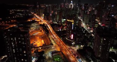 Gece: Yoğun, karanlık bir şehirde, gökdelenlerle çevrili parlak ışıklı bir yol yılanı..