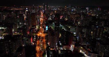 Gökdelenlerle çevrili yoğun ve aydınlık bir yolu gece vakti karanlık bir şehir manzarasından geçen bir hava görüntüsü yakalıyor..