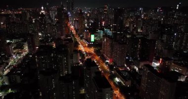 Gökdelenlerle çevrili yoğun ve aydınlık bir yolu gece vakti karanlık bir şehir manzarasından geçen bir hava görüntüsü yakalıyor..