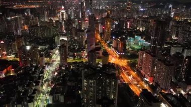 Gökdelenlerle çevrili yoğun ve aydınlık bir yolu gece vakti karanlık bir şehir manzarasından geçen bir hava görüntüsü yakalıyor..