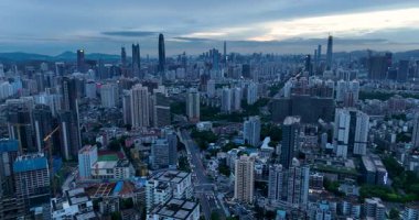 Alacakaranlıktaki yoğun, genişleyen şehir manzarası gökdelenleri ve bulutlu, karanlık bir gökyüzünün altındaki binaların arasından geçen ana yolu gösteriyor..