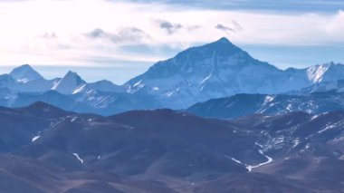 Mavi dağ katmanları kar yamasıyla aydınlık, kısmen bulutlu gökyüzünün altında geri çekiliyor..