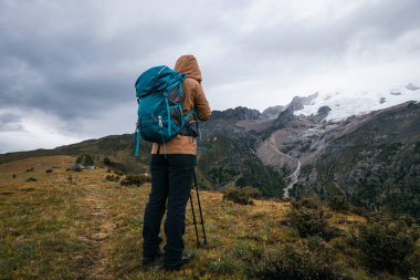 Sırt çantalı bir kadın yüksek irtifada dağ yürüyüşü yapıyor. Yağmurda karla kaplı dağa bakıyor.