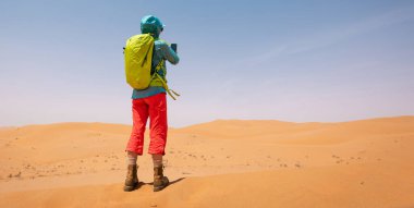 Sırt çantalı bir kadın çölde akıllı telefonuyla fotoğraf çekiyor.