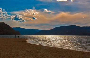 Gün batımında nehir kıyısında, parlak sularla, arka planda dağların siluetiyle....
