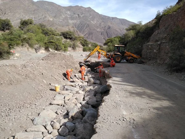 Yağmur mevsiminde nehirdeki su geçidinde çöken yola istikrar sağlayacak bir istinat duvarı inşa etmek için patinaj dümenine sahip adamlar.
