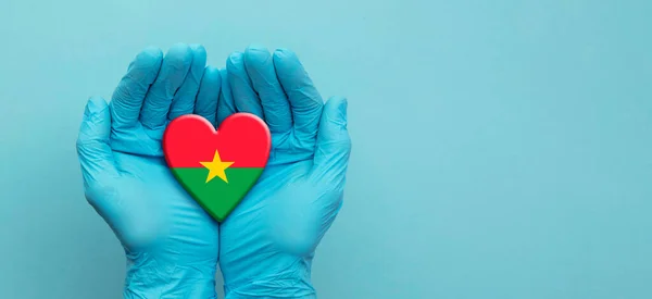 Medici mani che indossano guanti chirurgici tenendo il cuore bandiera Burkina Faso — Foto Stock