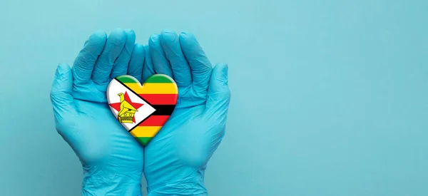 Medici mani che indossano guanti chirurgici che tengono il cuore bandiera Zimbabwe — Foto Stock