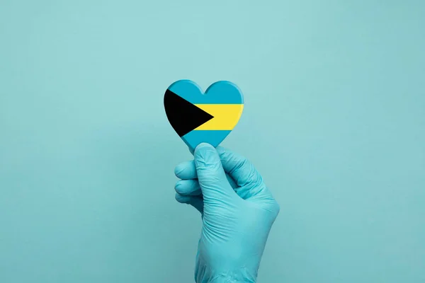 Mani che indossano guanti protettivi chirurgici che tengono il cuore bandiera Bahamas — Foto Stock