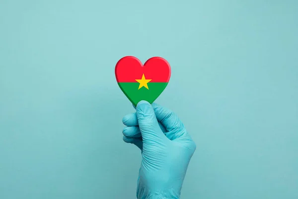 Manos con guantes quirúrgicos protectores que sostienen el corazón de la bandera de Burkina Faso — Foto de Stock