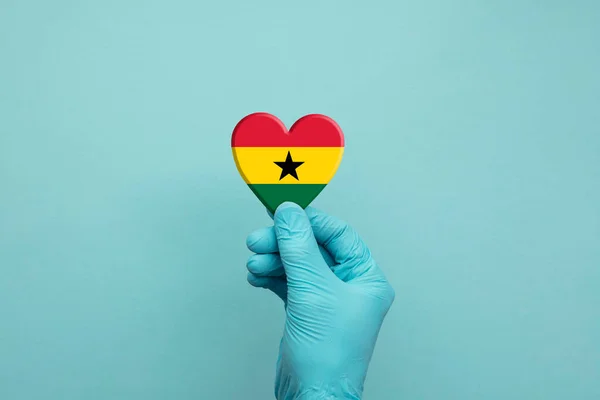 Mani che indossano guanti protettivi chirurgici che tengono il cuore bandiera Ghana — Foto Stock