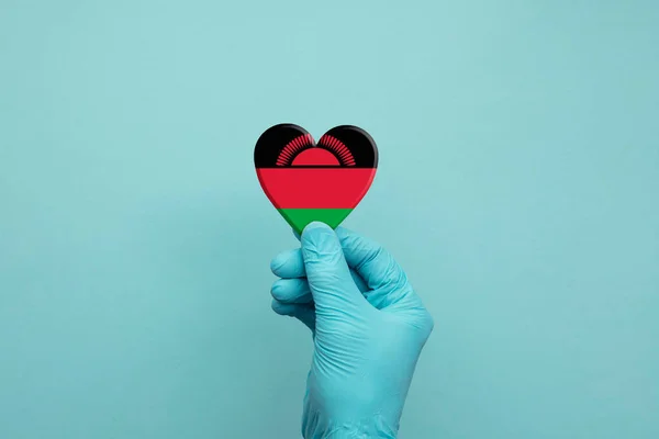 Mani che indossano guanti protettivi chirurgici che tengono il cuore bandiera del Malawi — Foto Stock