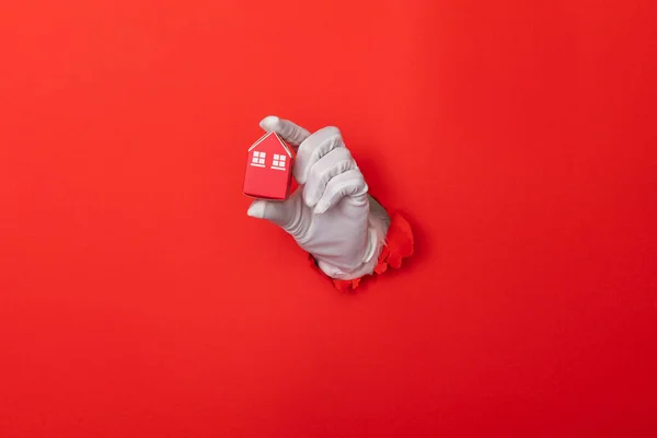 Santa Claus hand holding model house through a hole in red paper background — Stockfoto