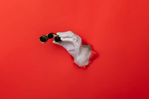 Santa Claus hand with binoculars through a hole in red paper background — Stockfoto