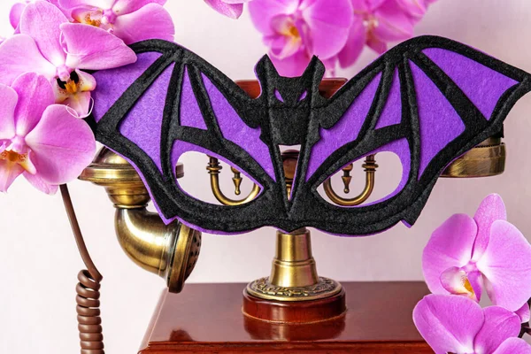 Bat shaped mask on vintage phone among pink orchid flowers