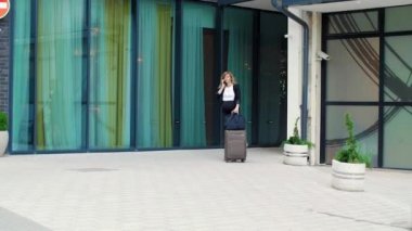 Modern binanın önünde bavulla yürüyen ve cep telefonundan bir şeyler okuyan gülümseyen hamile bir iş kadını. Arkaplan, perdeli büyük cam pencerelerden oluşur.