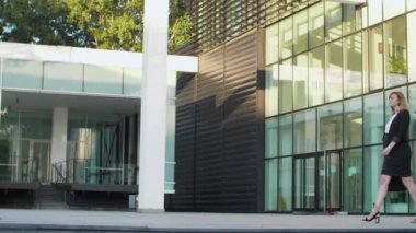 Hamile bir iş kadını modern bir binanın önünde yürüyor ve kol saatine bakıyor..