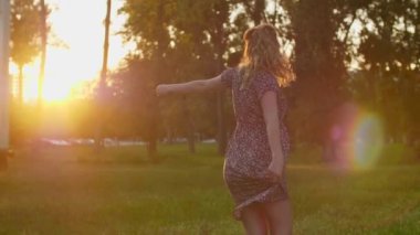 Günbatımında arka planda ağaçlarla şehir parkında duran gülümseyen beyaz hamile bir kadın. Elbisesini tek eliyle tutuyor..