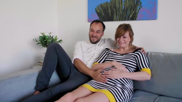 一对拥抱的年轻夫妇躺在客厅的沙发上 他们高兴地看着电视 那个男人一只手扶着她怀孕的肚子 — 图库视频影像