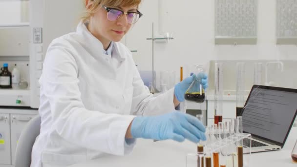 化学者は実験室で麻CbdとCbdaの油で働いています 彼女はガラス管とエルレンマイヤーのフラスコを使用して ラップトップで結果を書いています 大麻の種からの医療薬局 — ストック動画