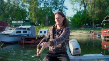 Beyaz orta yaşlı adam eski beyaz teknesinde kürek çekiyor. Gün doğumunda nehir marinasında. Bu balıkçı eski tahta küreği kullanıyor. Etrafında kayıklar ve yüzen evler ve arka planda ağaçlar var..