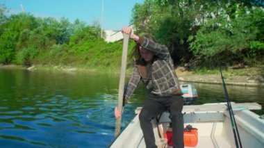 Mutlu balıkçı, günbatımında kürek çekmek için eski tahta kürek kullanıyor. Uzun kahverengi saçları var ve nehir marinasında beyaz bir teknede takılıyor. Etrafında ağaçları olan kayıklar ve yüzen evler var..