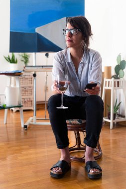 Genç bir bayan ressam ev stüdyosunda oturuyor ve pencereden içeri bakıyor. O bir bardak beyaz şarap ve cep telefonu tutuyor. Arka planda resim yapmak için gerekli malzemeler görülebilir..