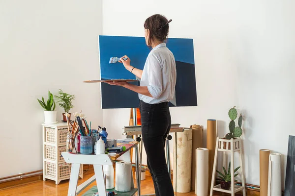 A young smiling painter applies paint to a canvas in her home studio. Other accompanying materials for painting could be seen around it. In some photos, she holds a palette of paints in one hand, while with the other she applies the paint with a pain