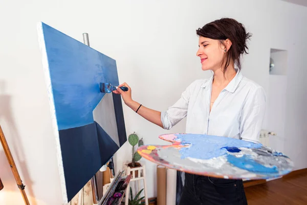 A young smiling painter applies paint to a canvas in her home studio. Other accompanying materials for painting could be seen around it. She holds a palette of paints in one hand, while with the other she applies the paint with a paintbrush on the ca