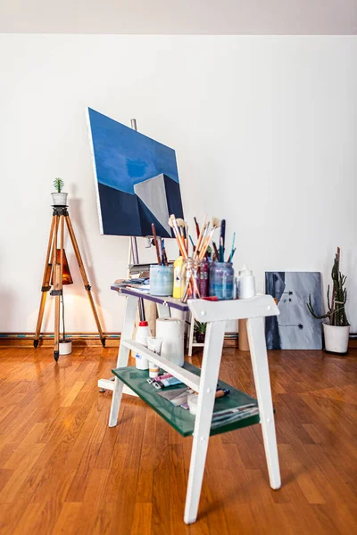 A painters work of art in studio. Around you can see a contemporary work of art placed on an easel, other pieces of art, as well as other accompanying material for painting.