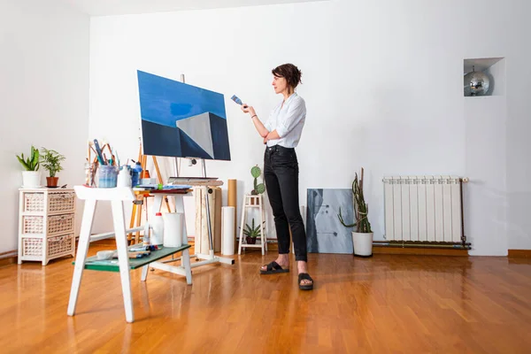 A young painter observes her work of art in her home studio. Around her, you can see a contemporary work of art placed on an easel, other pieces of art, as well as other accompanying material for painting.