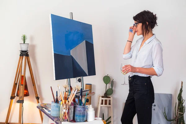 A young painter with a glass of wine stands in her studio and observes her work of art on canvas. Besides you can see around her other accompanying material for painting.