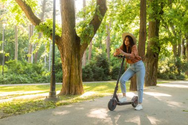 Telefonunda yazarken elektrikli scooterının yanında duran şapkalı güzel bir kadın. Arka planda ağaçlar ve yeşil renkler baskın.
