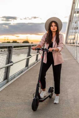 Genç ve güzel bir kadın bir ayağı ayakta duran bir platformda elektrikli scooterının yanında duruyor. O bir şapka takıyor ve iş eğitimli. Arkaplan bir köprünün parçalarından oluşur