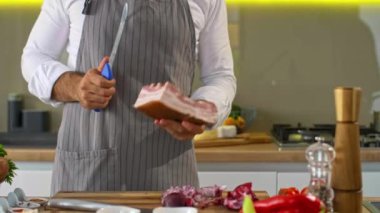 Şef ahşap bir tahtanın üzerinde mutfakta domuz pastırması keser. İş yüzeyinin etrafında sebzeler, ekmek dilimleri, baharatlar ve küçük mutfak kaseleri var..
