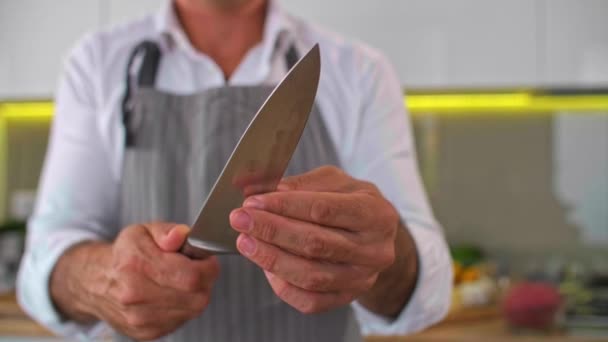 シェフは台所で食事を準備する前にナイフを研ぎます 前景には両手と野菜 スパイス 小さなキッチンボウルが描かれています — ストック動画