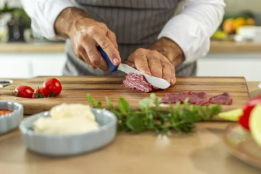 Ahşap mutfak tahtasının hazırlık safhasında şefin ellerinin carpaccio etini ince parçalara ayırması. Etrafta çeşitli küçük mutfak kaseleri ve sebzeler görülebilir..