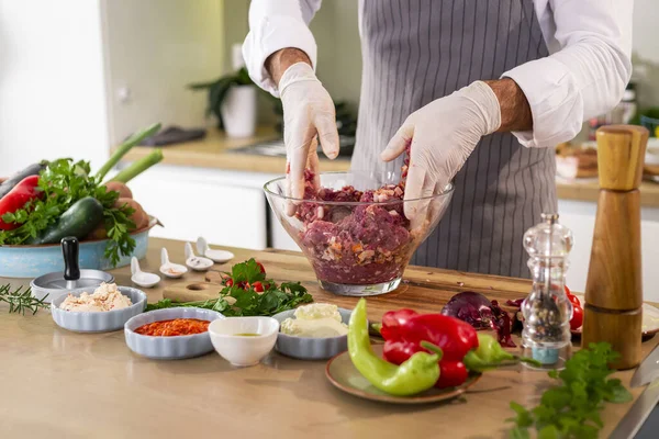 The chef mixing meatballs with spices and vegetables in a transparent glass bowl, while on the side of the work surface there are vegetables, slices of bacon, spices and small kitchen utensils.