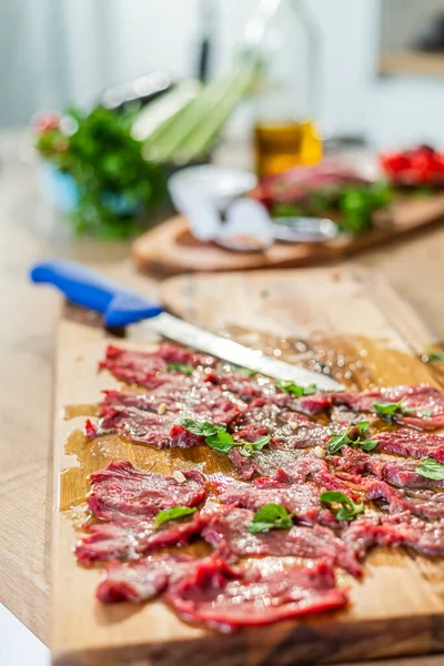 Close-up display on a wooden kitchen board of ready-made carpaccio meat for tasting. Various vegetables can be seen around, as well as small kitchen bowls with side dishes.
