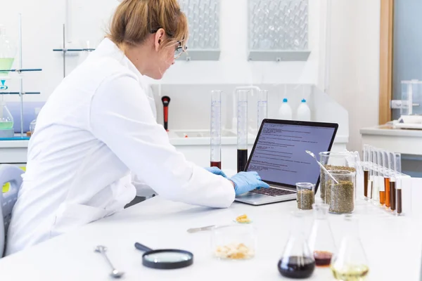 Laboratuvarda dizüstü bilgisayarda çalışan kadın kimyacı geniş açıdan görülüyor. Kenevir tohumu, CBD ve CBDA cbd yağları masada. Sağlık hizmetleri eczanesi esrar kavramı.