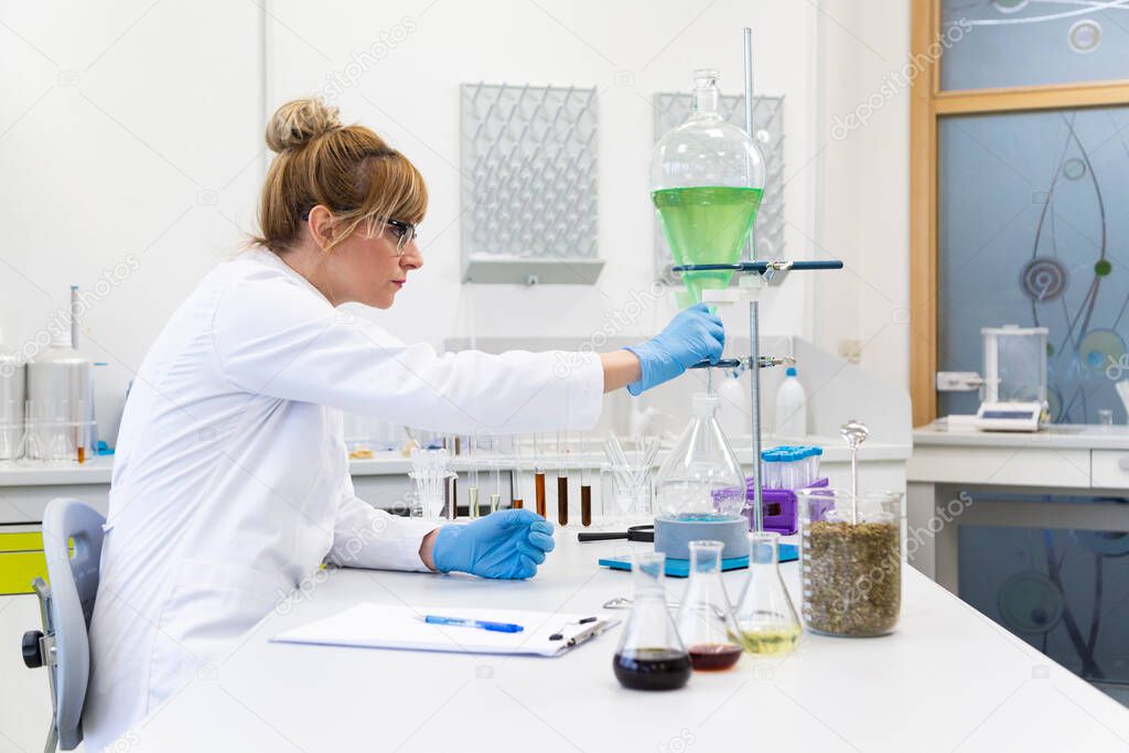 White female scientist wearing glasses and latex gloves works with green turquoise liquid in pear shaped separator funnel. Hemp seeds and cbd oils are on table.