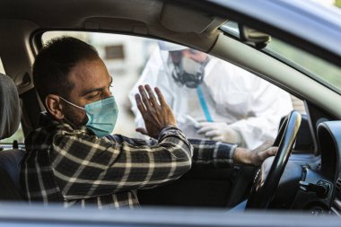 Adam sürüş için COVID 19 testi yapmayı reddediyor, hastadan araba camından burun örneği alıyor, PCR teşhisi koyuyor, PPE 'de elinde test kiti olan bir doktor..