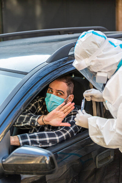 Man refuses medical worker trying to perform drive-thru COVID-19 test, taking nasal swab sample from patient through car window, PCR diagnostic, doctor in PPE holding test kit. She is holding passports.