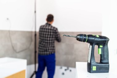 Matkap dairenin odasındaki çalışma yüzeyine yerleştirilir, arka planda ise rafları yerleştirmek için duvarı delme işini yapan bir marangoz görebilirsiniz..