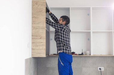 Marangoz vidaları vidalıyor ve mutfak parçalarını elektrikli tornavidayla birleştiriyor. Etrafında iş aletleri ve çalıştığı malzemeler var..