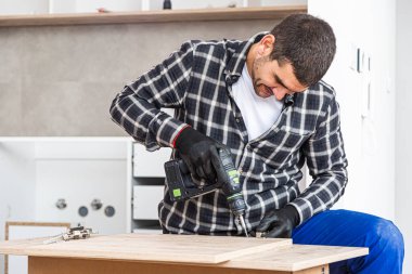 Marangoz vidaları vidalıyor ve mutfak parçalarını elektrikli tornavidayla birleştiriyor. Etrafında iş aletleri ve çalıştığı malzemeler var..