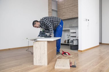 Bir marangoz odanın zemininde çalışma yüzeyinin ahşap bir tahtasını kesiyor. İşini, kendisine bir çalışma masası olarak hizmet eden ahşap bir kutuda yapar..