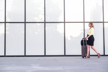 Hamile, beyaz bir kadın, bir parça seyahat bagajıyla modern bir binanın önünden geçiyor. Arkaplan büyük bir cam pencere içeriyor.