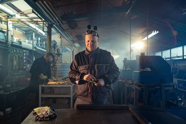 Tamirci, otomatik kaynak aletini işe hazırlarken meslektaşı arkada öğütme işiyle uğraşırken fark edilebilir..